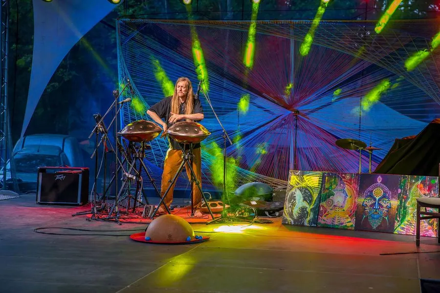 David Lesage seul sur la scène du Everness Festival, en Hongrie : debout pieds nus derrière ses deux handpans acoustiques montés sur des trépieds noirs, sous des faisceaux de lumière verte et rouge, devant une grande toile tissée multicolore ; une demi-sphère de calebasse est posée au sol devant lui.