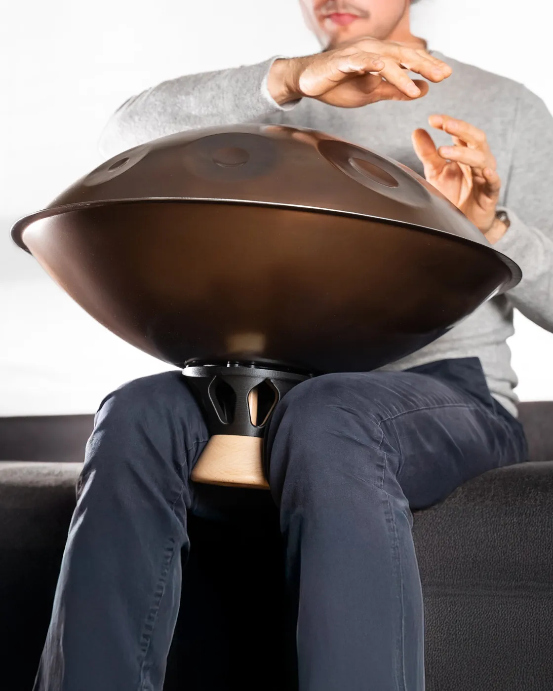 Une personne assise sur un canapé joue d’un handpan tenu par l’Atlas Body coincé entre ses cuisses : l’instrument est décollé de ses jambes.
