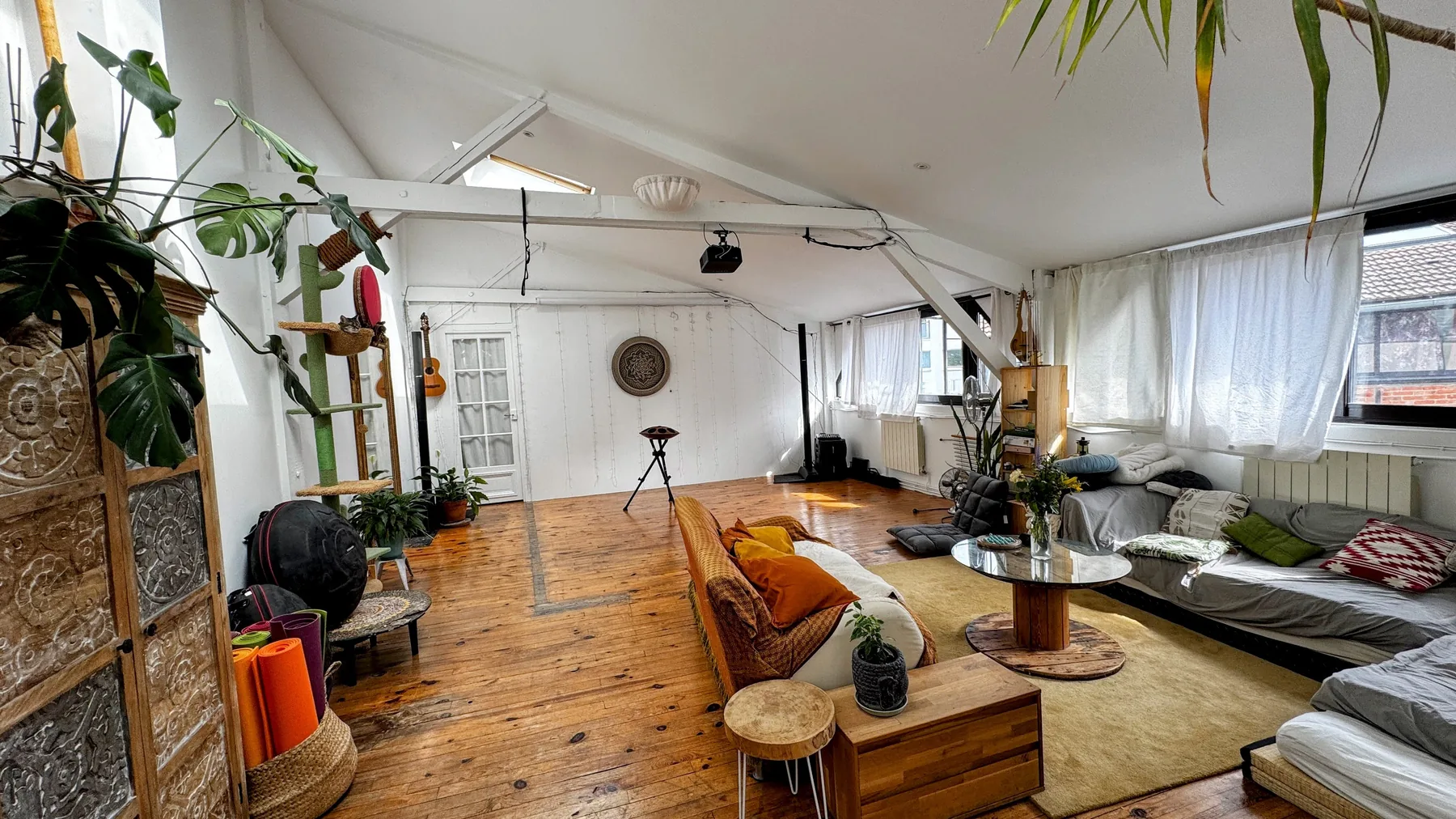 La grande pièce du showroom en journée, avant l’arrivée du public : parquet ancien, canapé bas et coussins posés à même le sol, un handpan acoustique sur son trépied au fond, une guitare accrochée au mur, des plantes et des tapis de sol roulés dans un panier.