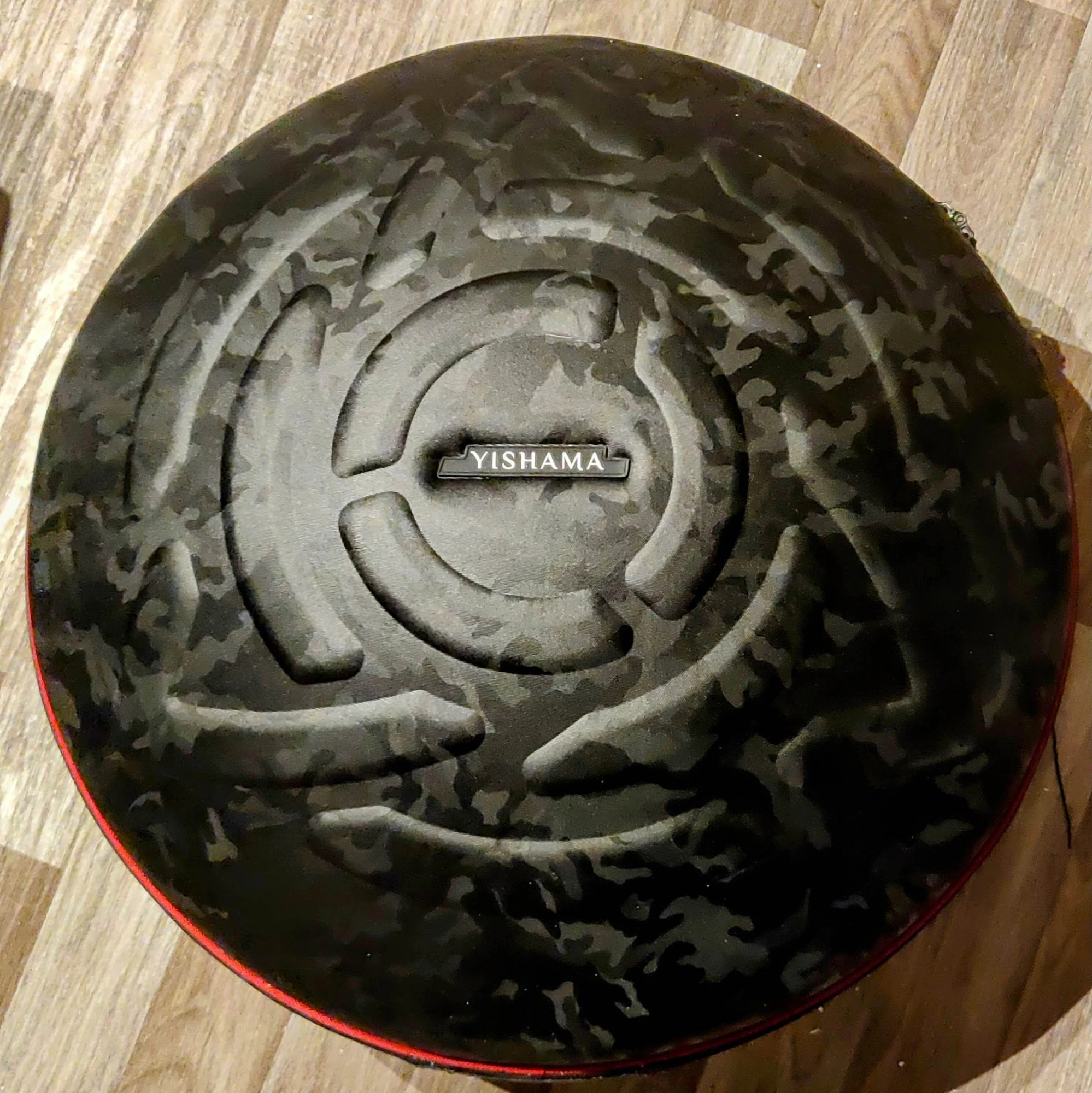 La housse rigide du handpan vue de dessus, posée sur un parquet clair : tissu camouflage noir mat, large spirale matelassée en relief, badge rectangulaire portant le mot YISHAMA au centre, liseré rouge sur la tranche et fermeture éclair sur le pourtour.