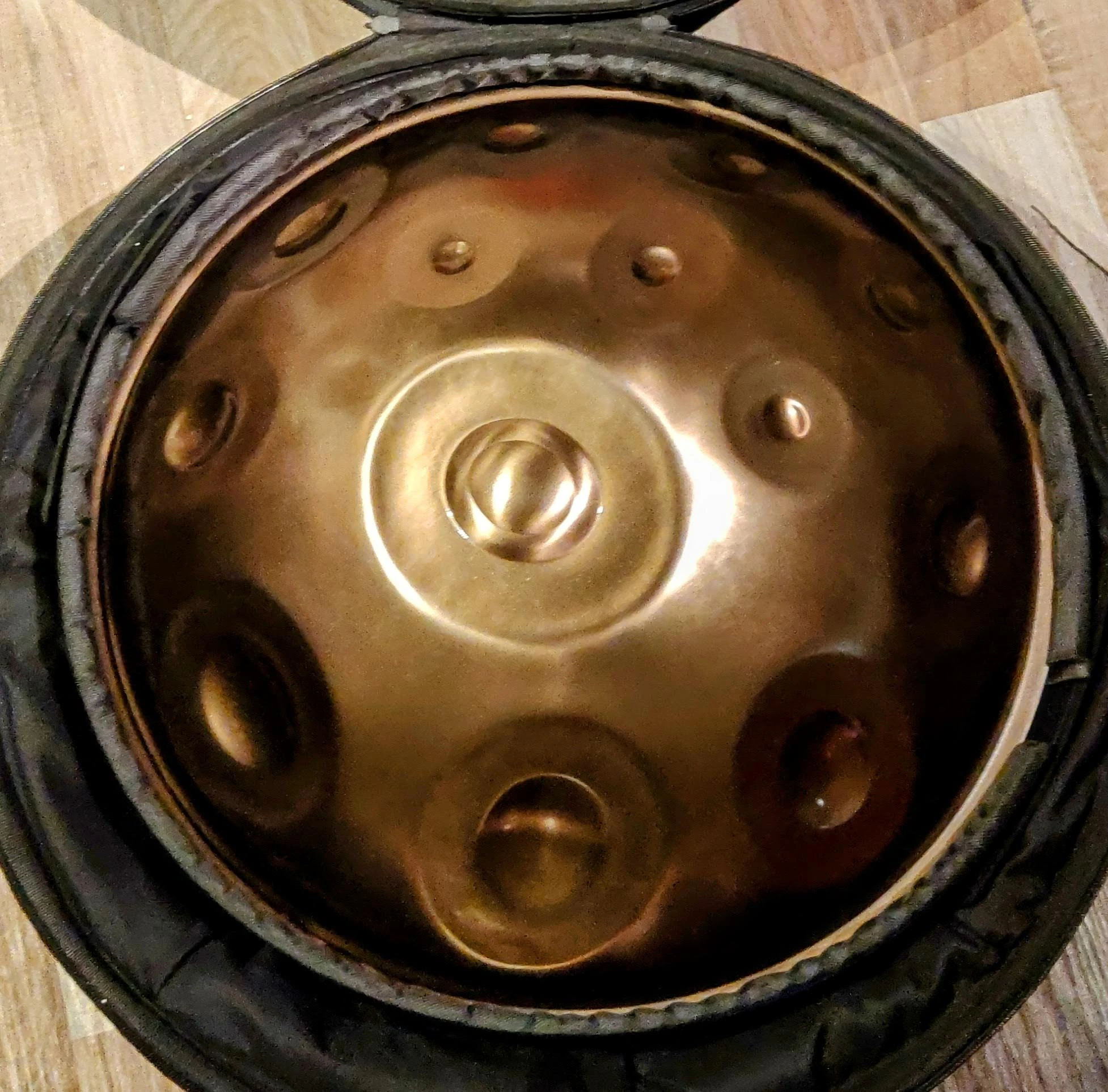 La housse ouverte : à l’intérieur, le handpan dont le métal renvoie une lumière chaude, dorée et bronze. Au centre, le ding et ses anneaux concentriques ; tout autour, les zones de notes, en creux et en dômes.