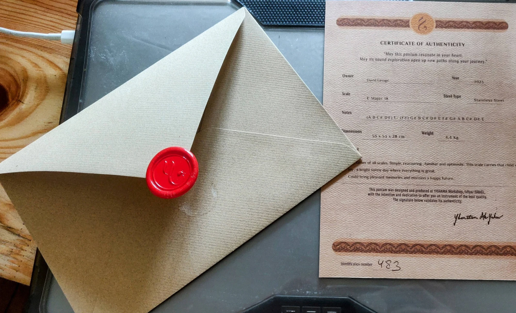 Sur un plan de travail en bois : l’enveloppe kraft, maintenant ouverte, avec son cachet de cire rouge frappé d’un monogramme, posée à côté du certificat d’authenticité imprimé sur papier texturé.