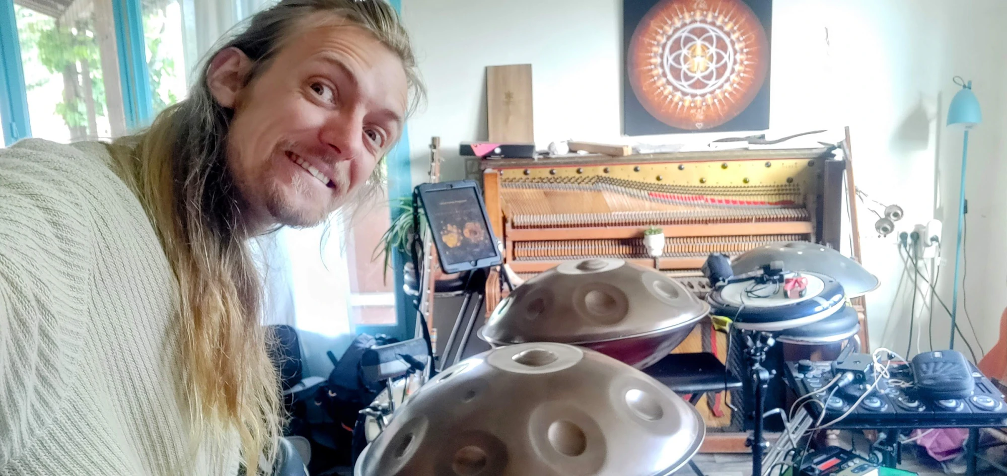 David Lesage souriant, tête penchée, en pleine lumière du jour. Devant lui, ses deux handpans Yishama en métal gris anthracite posés sur leurs pieds ; derrière, une tablette fixée sur un bras articulé, le cadre d’un piano droit ouvert laissant voir cordes et mécanique, un tableau représentant une Fleur de Vie orangée, des pads électroniques et un pédalier.